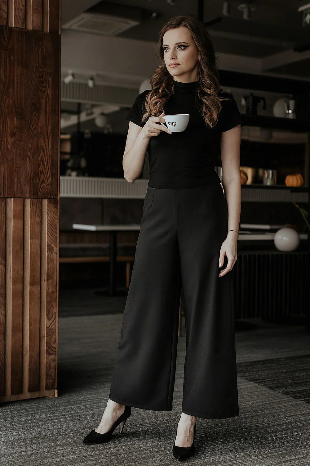 Woman in black wide-leg trousers, a black top and heels holding a coffee cup fashion image